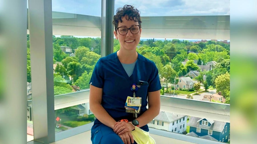 Erin Sabo sits in front of a window overlooking the University of Rochester Medical Center campus, wearing navy blue scrubs.